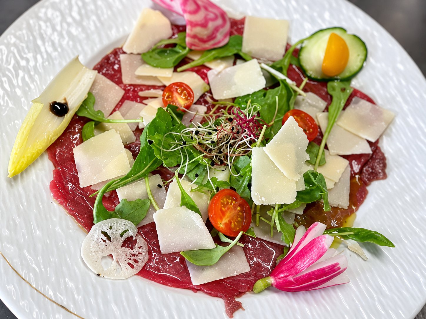 Assiette blanche de carpaccio de bœuf garnie de copeaux de fromage, de roquette, de pousses, de tomates cerises, de radis et d’une demi-endive.
