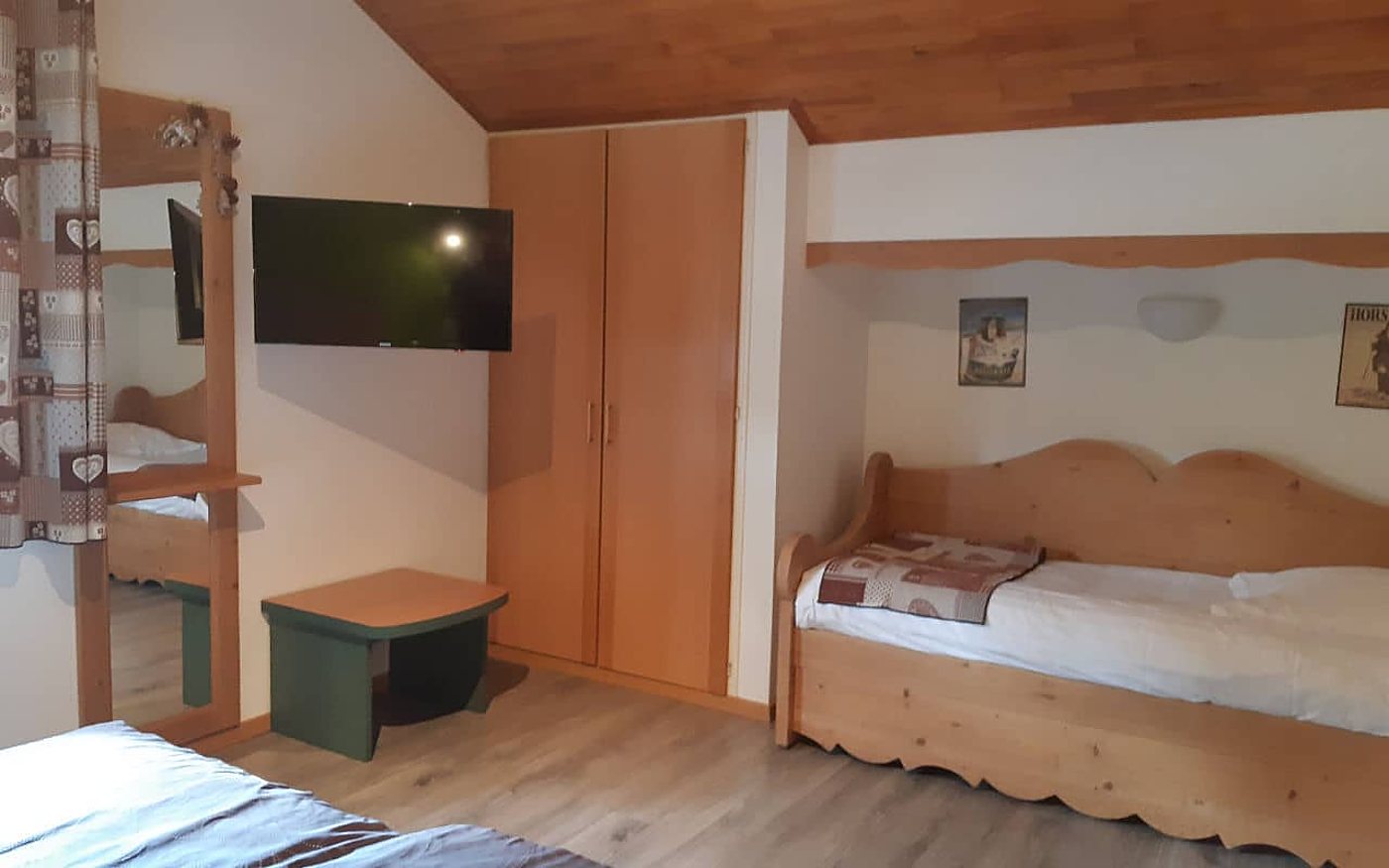 Chambre de style chalet avec lit banquette en bois clair, armoire, télévision murale, miroir et petit tabouret vert.