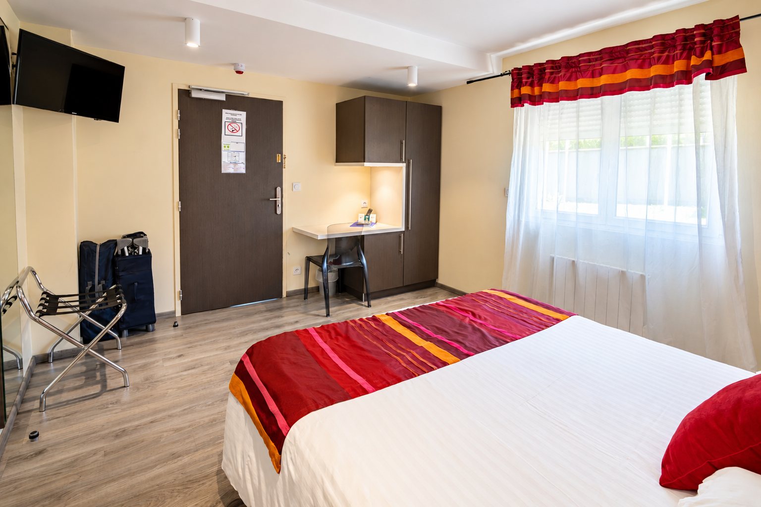 Chambre d’hôtel lumineuse avec un grand lit à plaid rouge et orange, un porte-bagages, un petit bureau, une télévision murale et une fenêtre à voilage blanc.