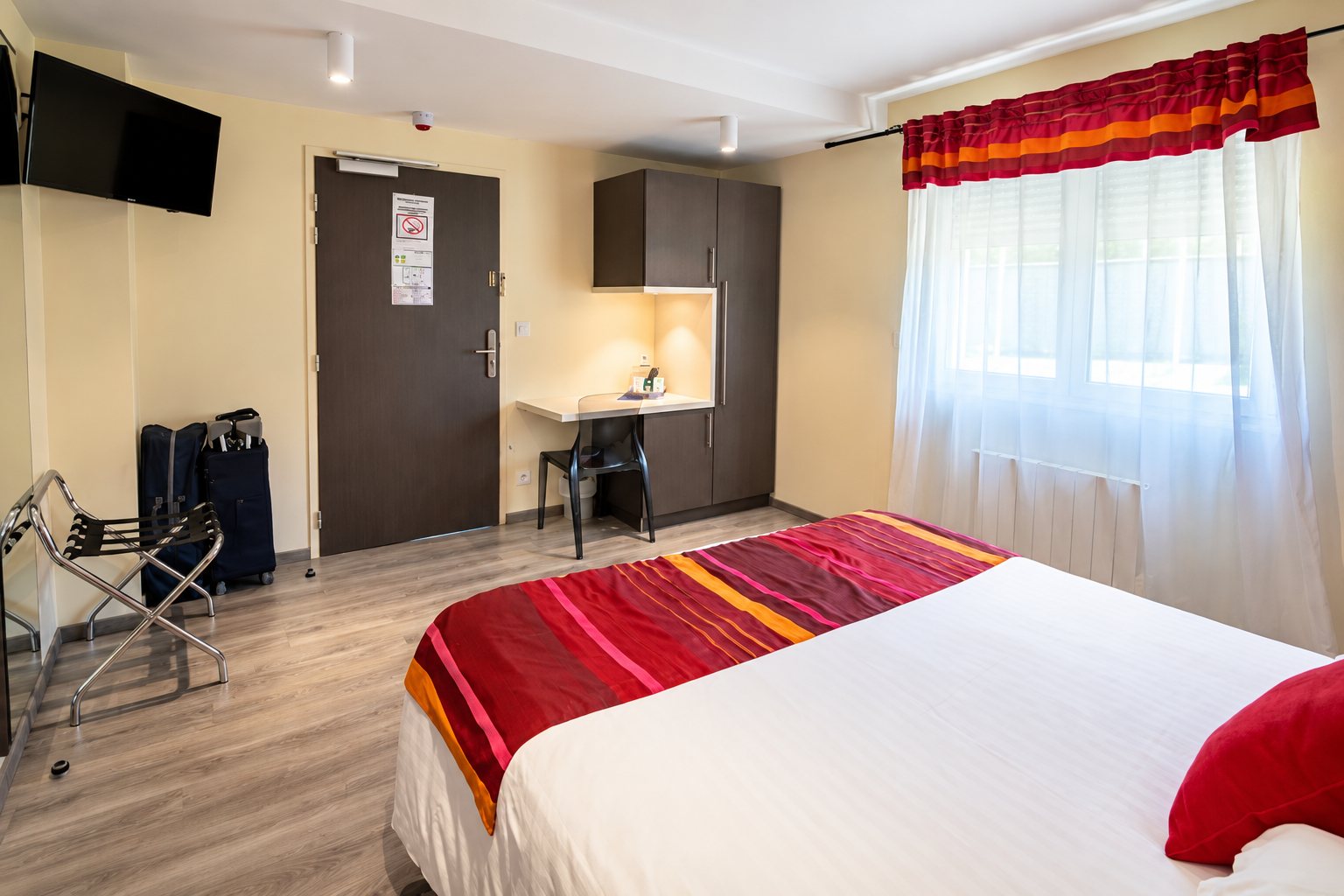 Chambre d’hôtel lumineuse avec un grand lit double à plaid rayé rouge et orange, un petit bureau, une télévision murale et des valises près de la porte.