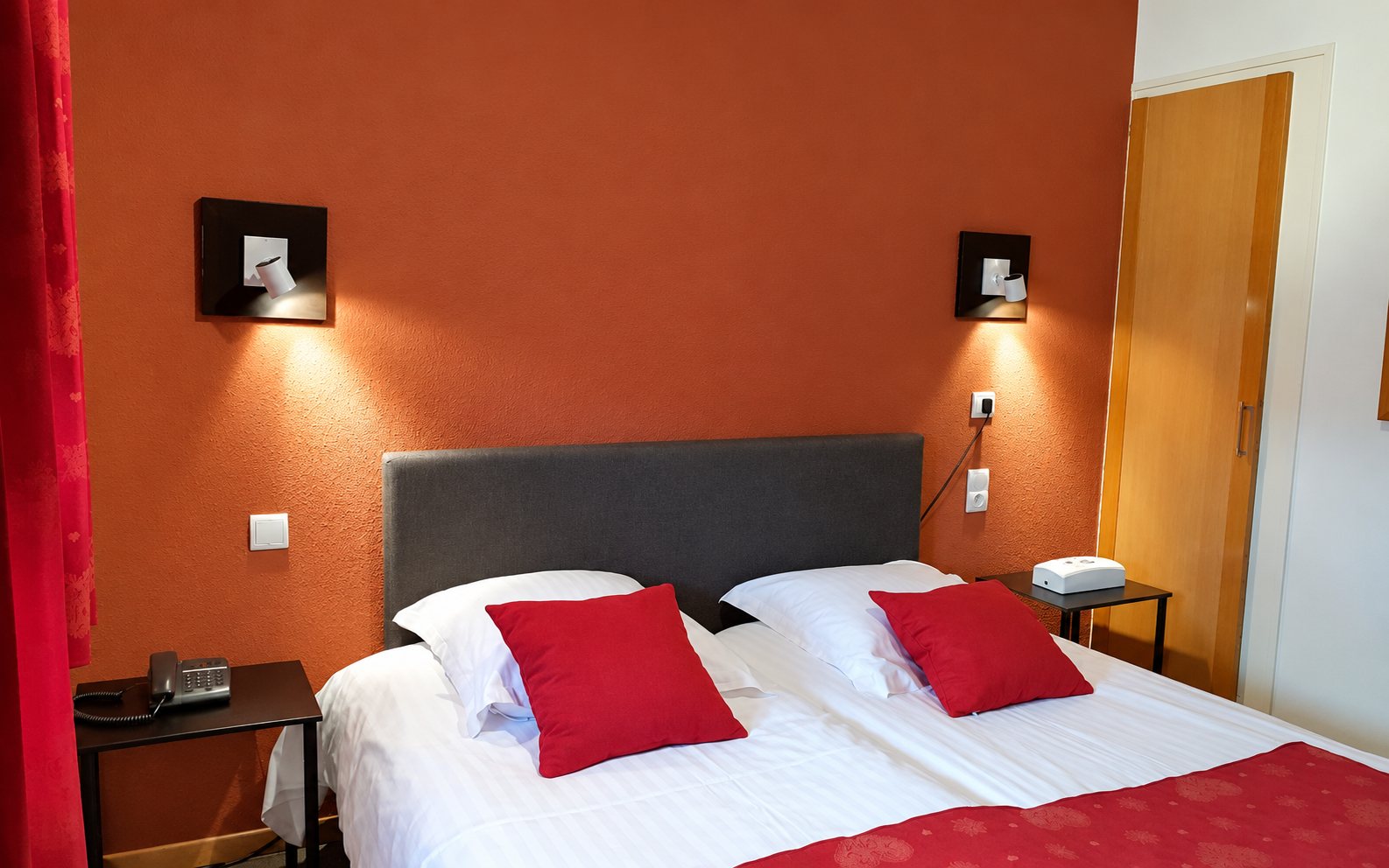 Chambre d’hôtel chaleureuse avec un lit double aux draps blancs, deux coussins rouges, deux appliques allumées et un mur orangé.