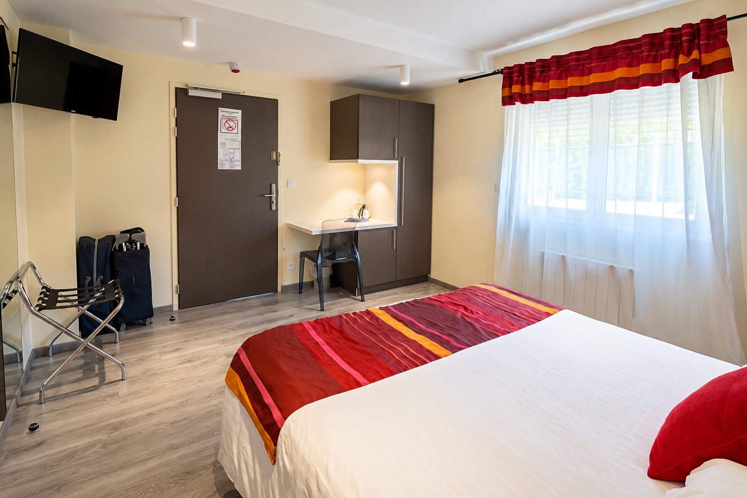 Chambre d’hôtel avec un grand lit double, un chemin de lit rouge et orange, un bureau, une télévision murale et des valises près de la porte.