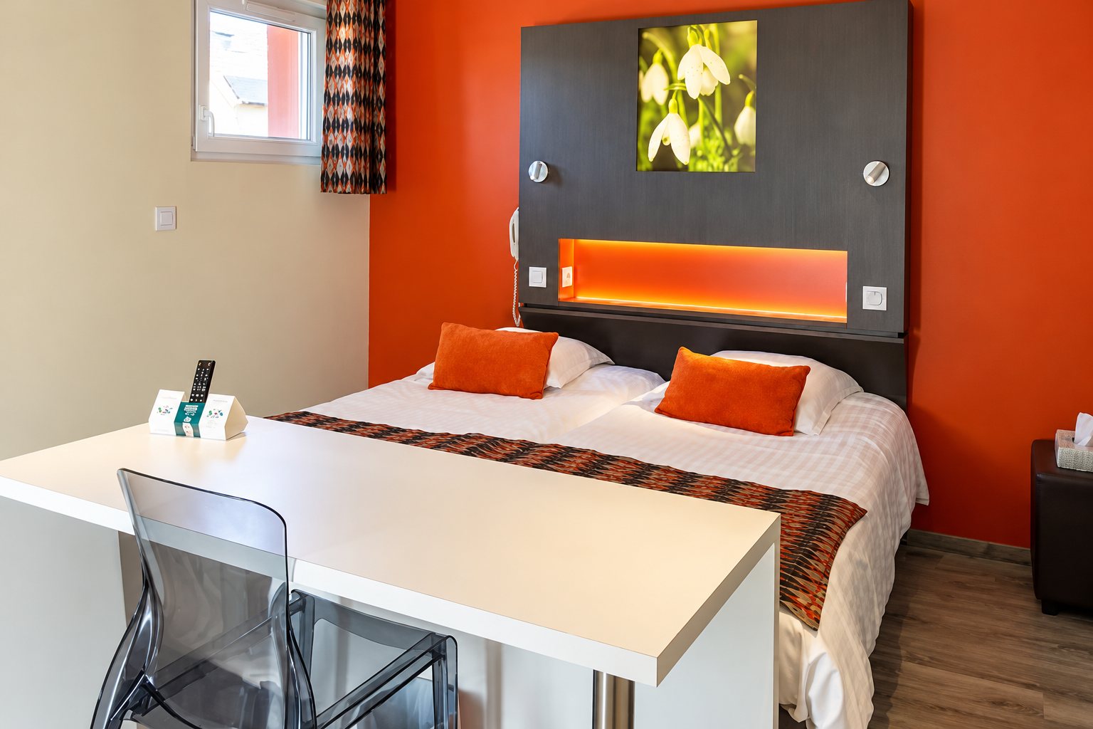 Chambre d’hôtel lumineuse avec un grand lit double, des coussins orange, un mur orange et un bureau blanc au premier plan.