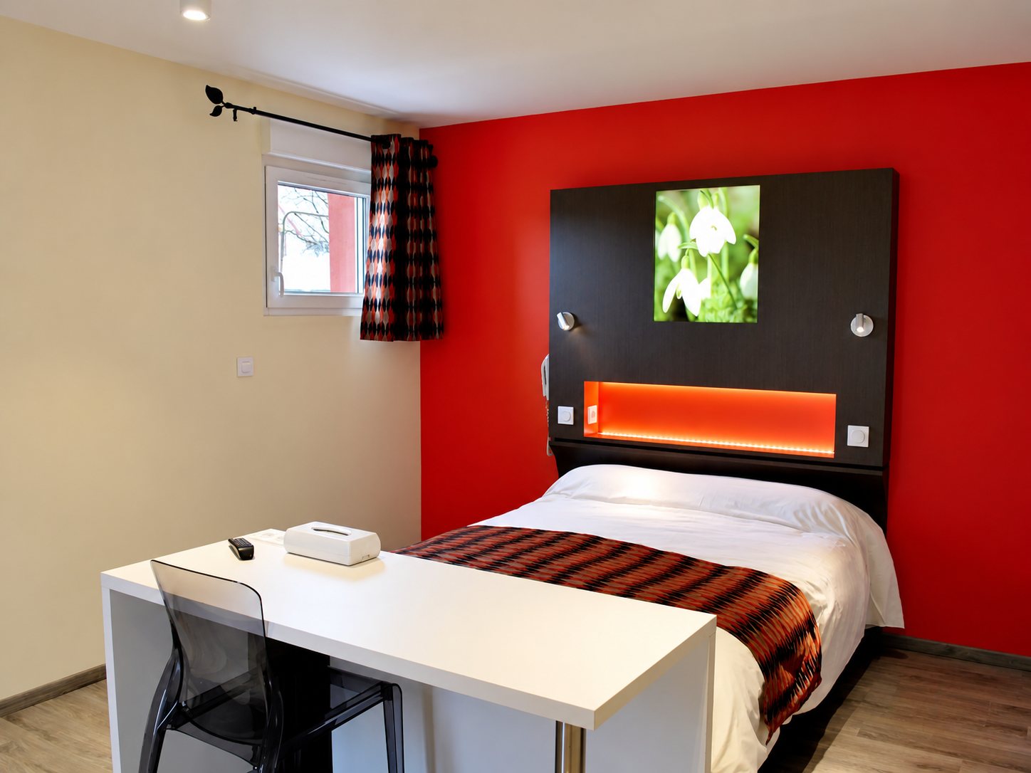Chambre d’hôtel moderne avec un mur rouge, un lit double à tête de lit sombre éclairée et un bureau blanc au premier plan.
