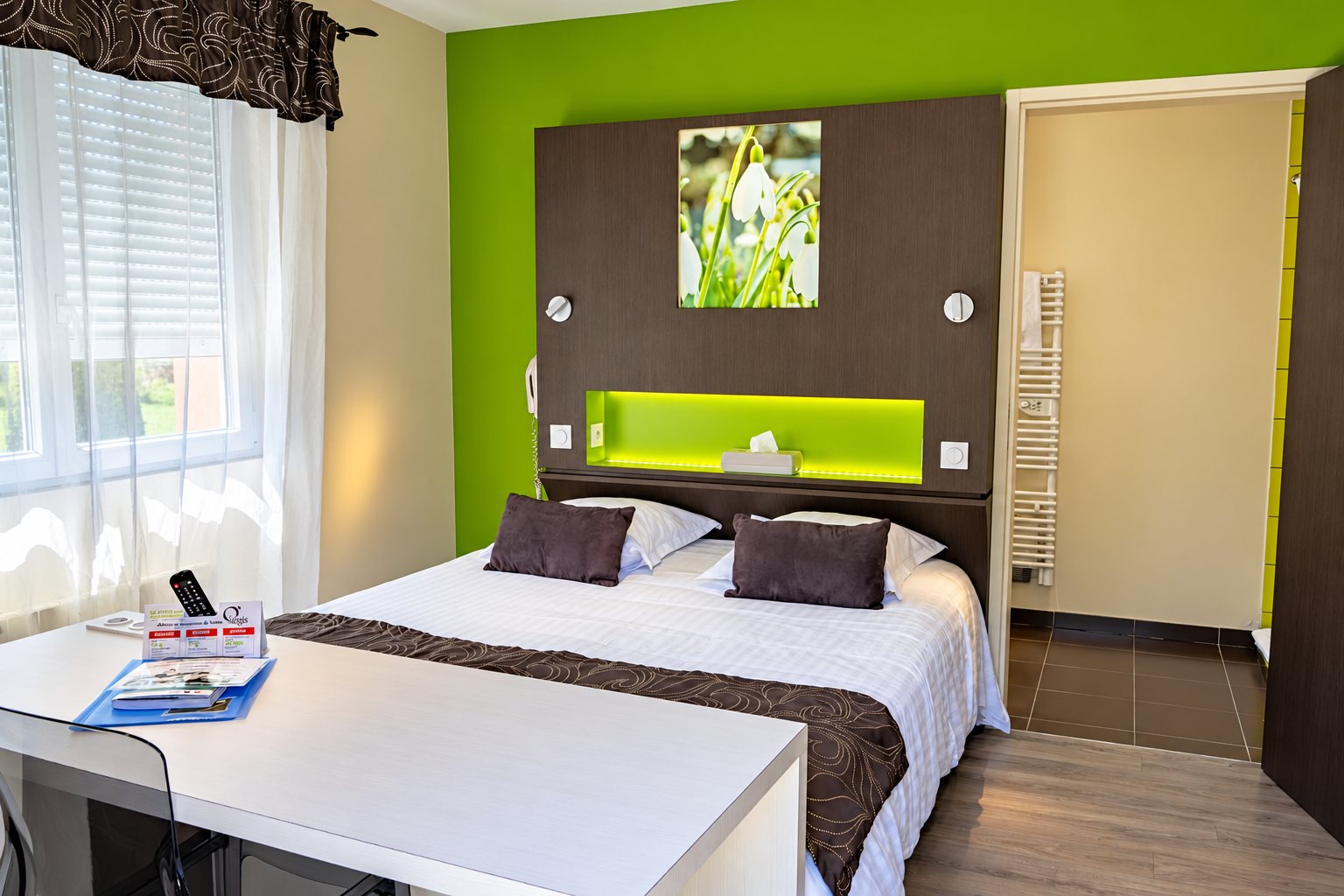 Chambre d’hôtel lumineuse avec un mur vert anis, un lit double à tête de lit grise ornée d’une photo de perce-neige, un bureau au premier plan et une salle de bain en arrière-plan.