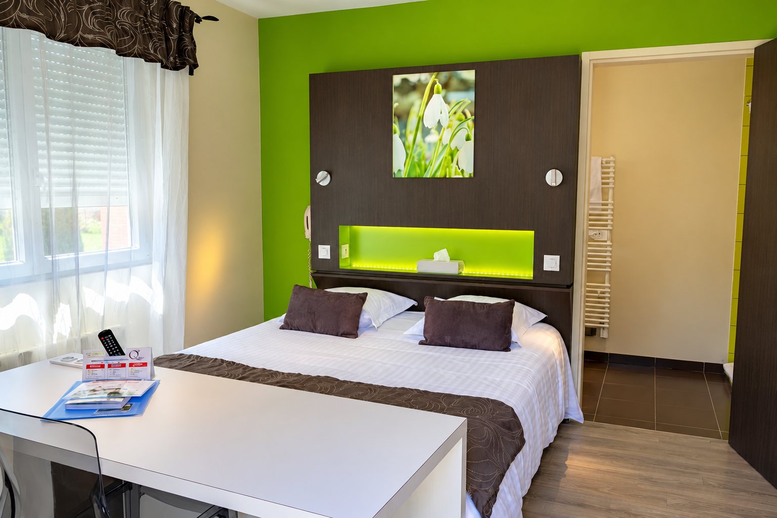 Chambre d’hôtel lumineuse avec un mur vert anis, un grand lit double aux coussins gris, un bureau blanc au premier plan et une salle de bain en arrière-plan.