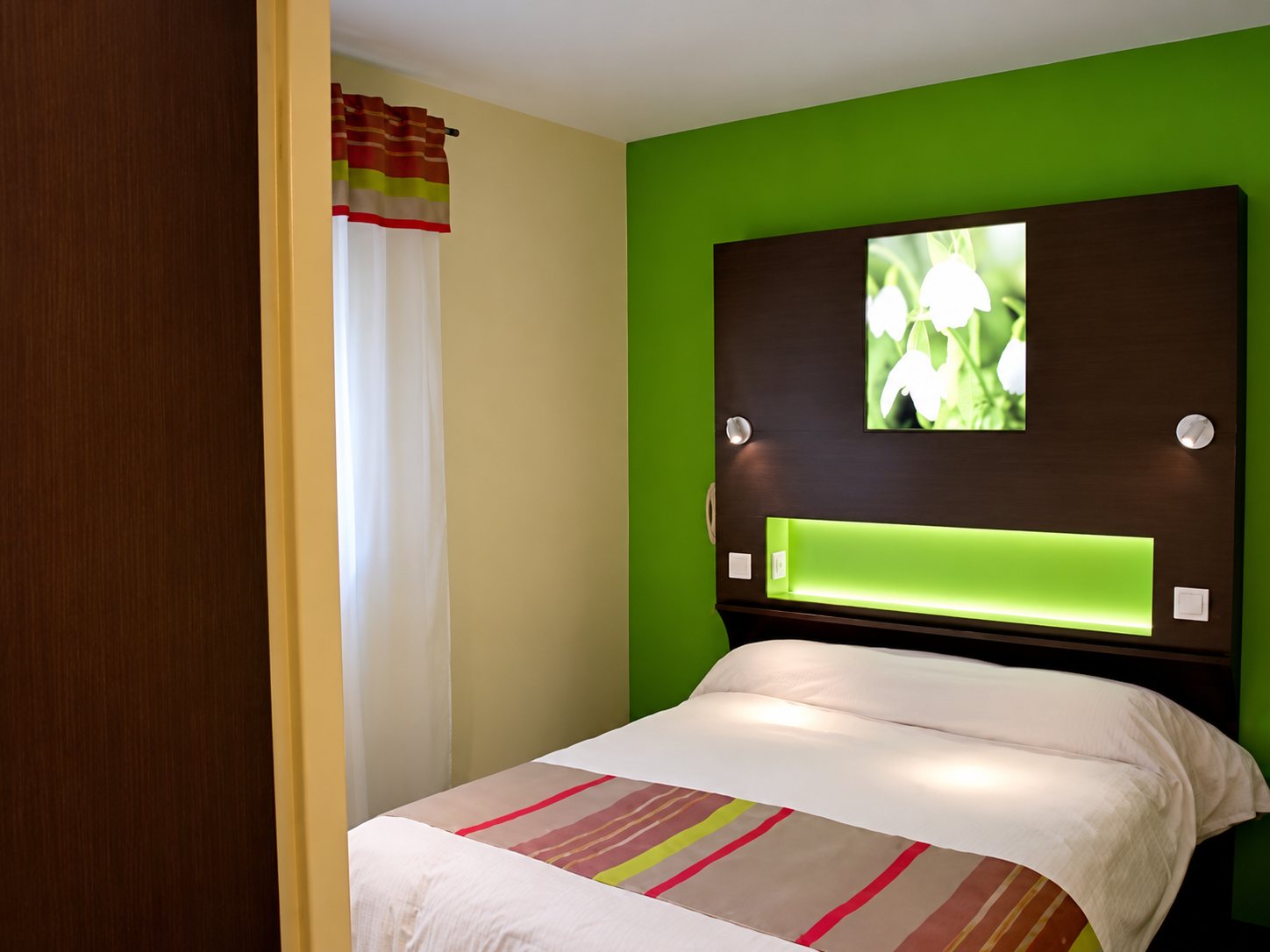 Une chambre d’hôtel avec un lit double, un mur vert, une tête de lit sombre éclairée en vert et un tableau de fleurs blanches.