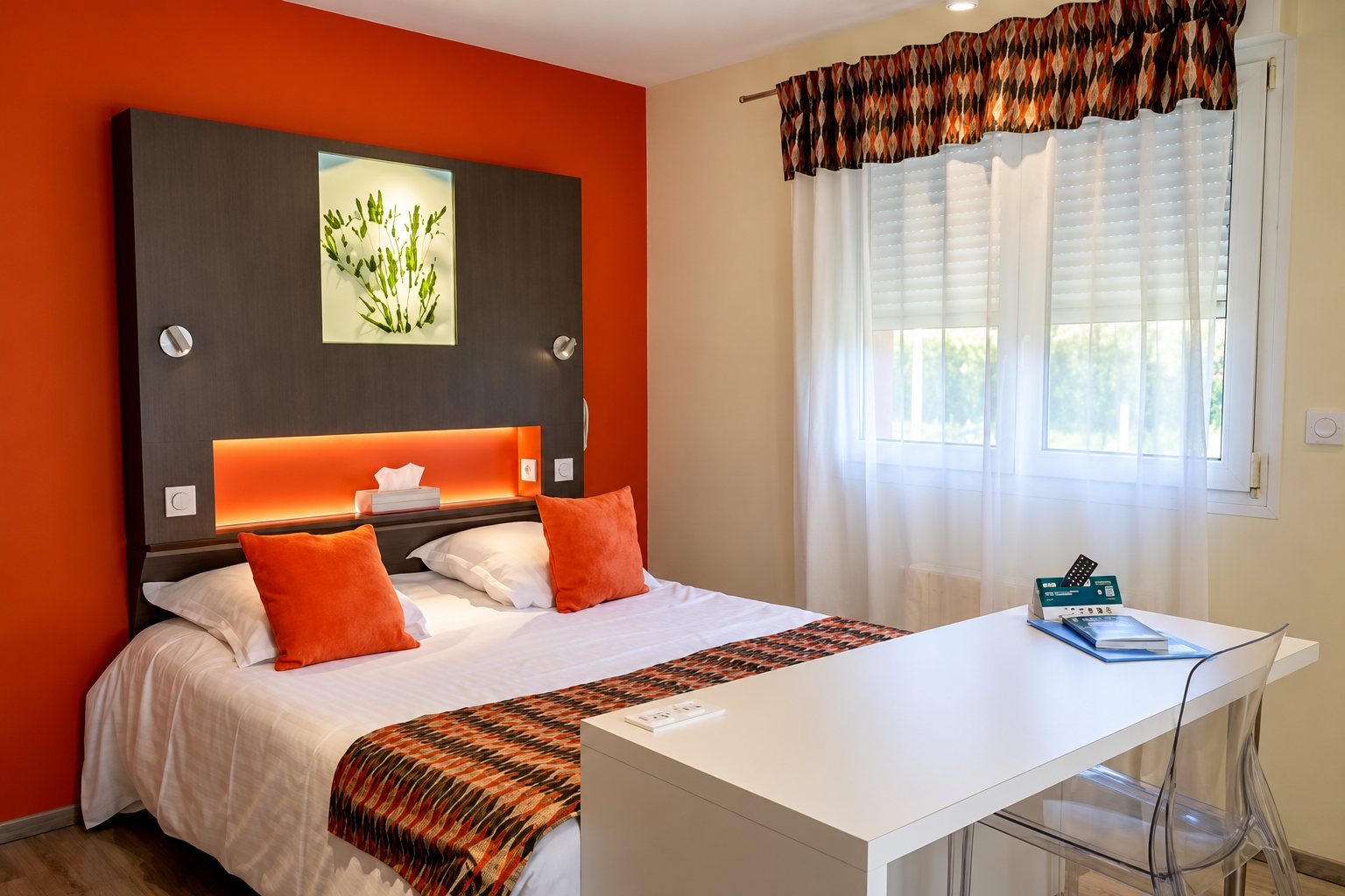 Chambre d’hôtel lumineuse avec un mur orange, un lit double aux coussins orange, un bureau blanc et une fenêtre aux rideaux voilés.