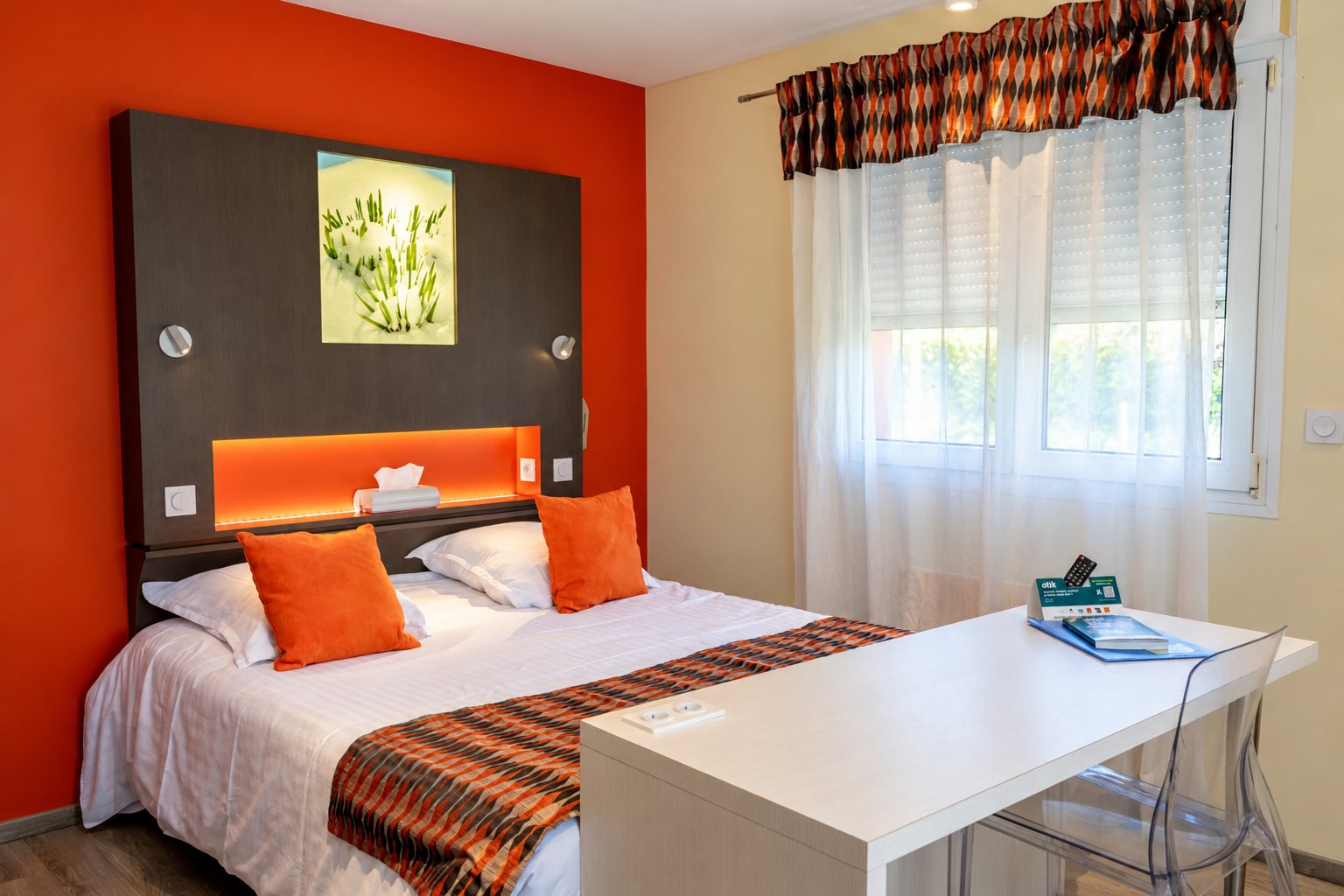 Chambre d’hôtel lumineuse avec un mur orange, un lit double aux coussins orange, un bureau blanc et une fenêtre aux voilages blancs.