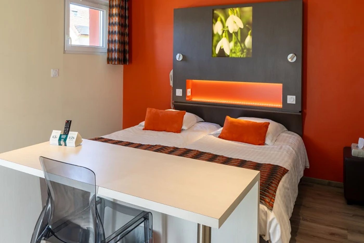Chambre d’hôtel moderne avec un mur orange, un lit double orné de coussins orange, une tête de lit grise éclairée et un bureau blanc au premier plan.