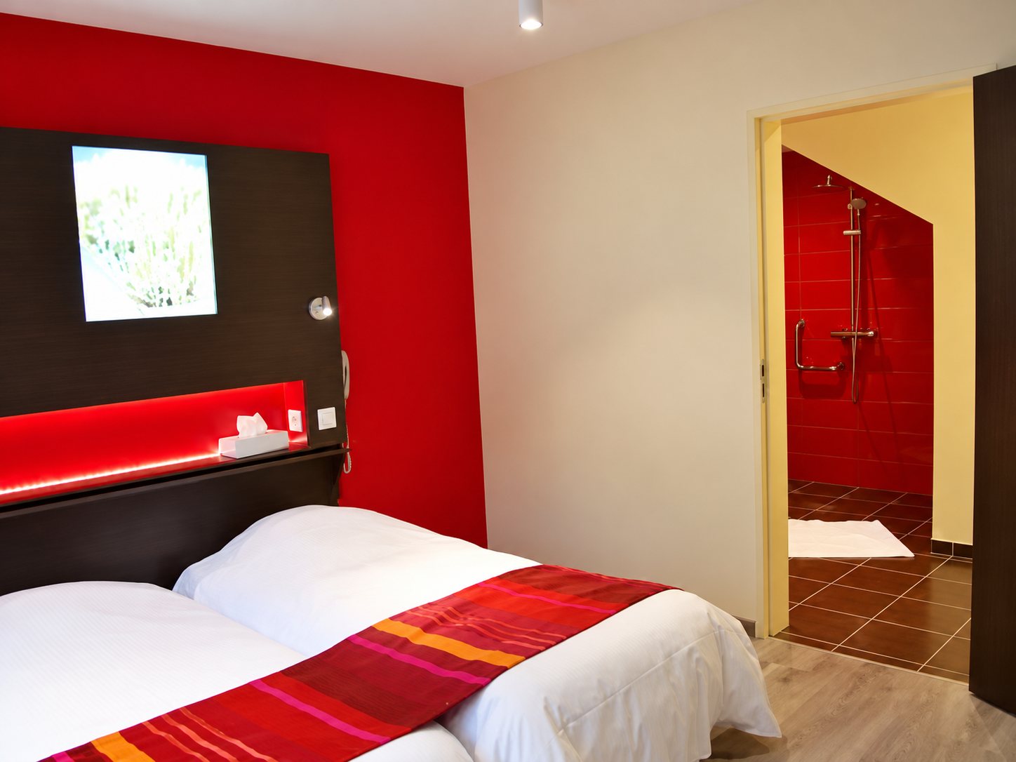 Chambre d’hôtel avec deux lits blancs ornés de chemins de lit rouges, un mur rouge et une salle de douche carrelée de rouge visible par la porte ouverte.