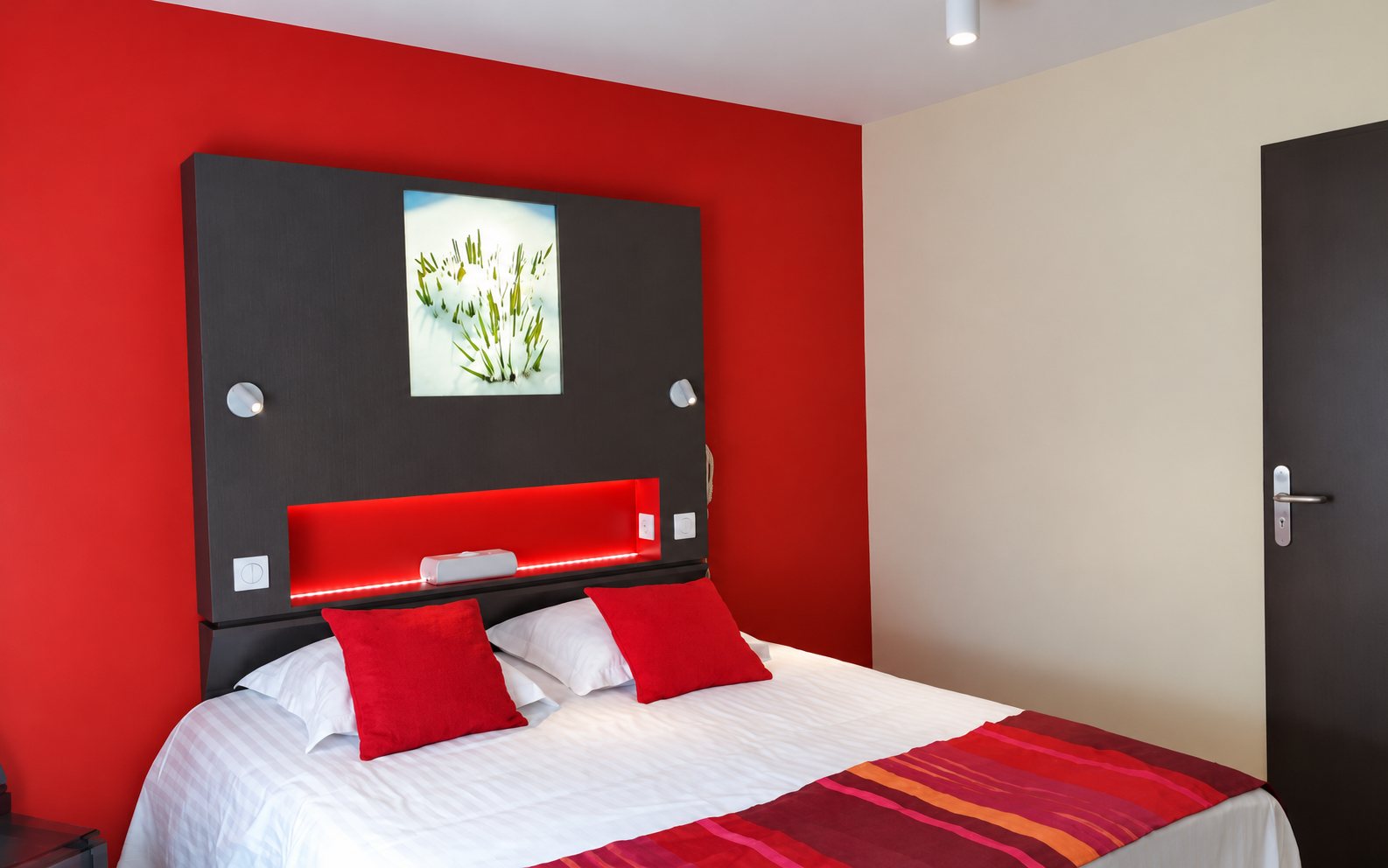 Chambre d’hôtel avec un lit double aux coussins rouges, une tête de lit grise éclairée en rouge et un mur rouge.