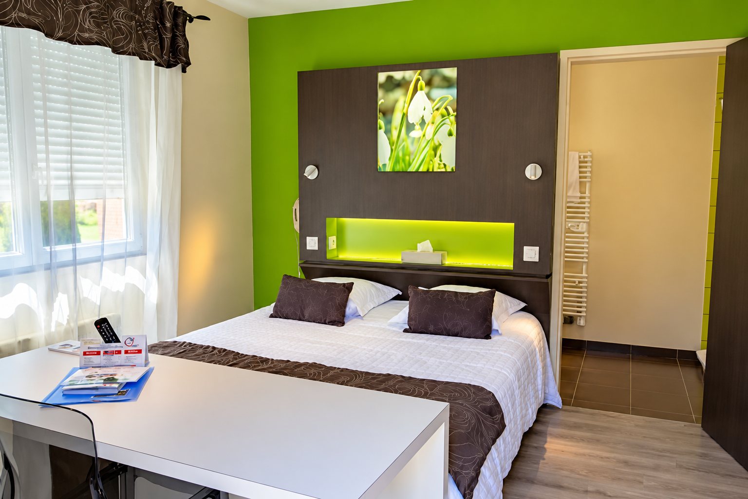 Chambre d’hôtel lumineuse avec un mur vert anis, un grand lit double aux coussins bruns, un bureau blanc au premier plan et une salle de bains en arrière-plan.