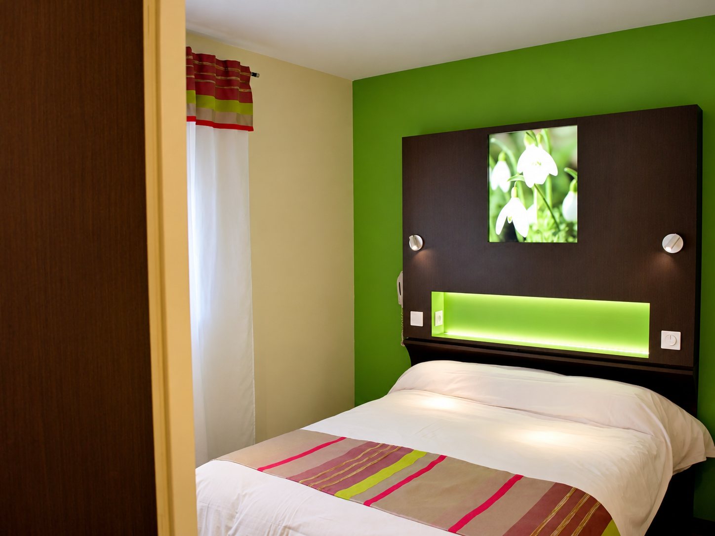 Chambre d’hôtel moderne avec un mur vert, une tête de lit sombre illuminée ornée d’une photo de perce-neige et un lit double avec chemin de lit rayé.