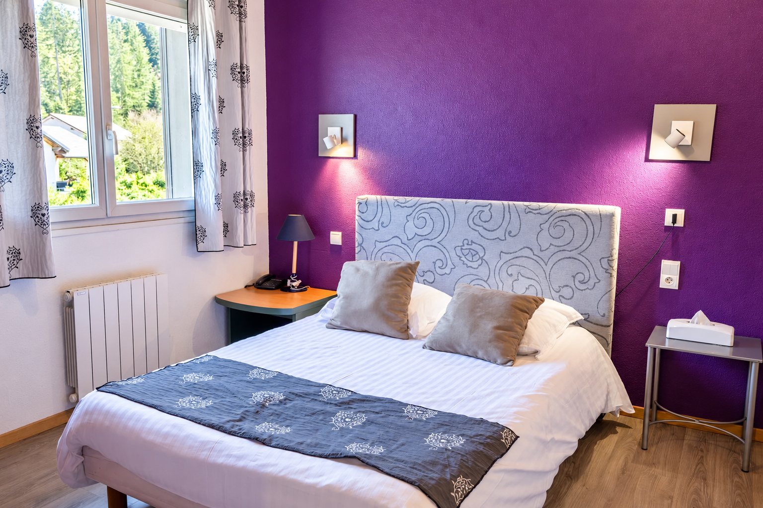 Chambre d’hôtel avec un mur violet, un lit double à tête de lit brodée, deux tables de chevet et une fenêtre donnant sur la verdure.
