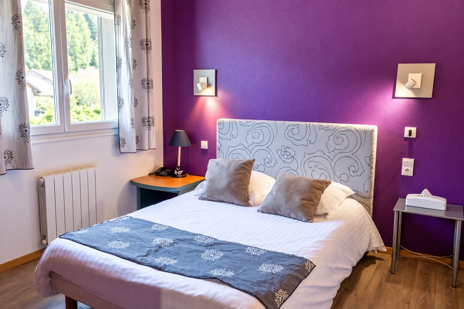 Chambre d’hôtel lumineuse avec un mur violet, un grand lit double aux coussins gris, une tête de lit décorée, une fenêtre et un radiateur blanc.