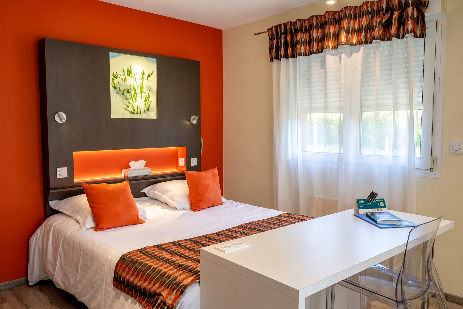 Chambre d’hôtel lumineuse aux murs orange avec un grand lit à coussins orange, une tête de lit grise éclairée, un bureau blanc et une chaise transparente.