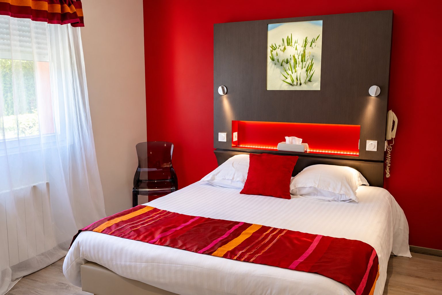 Chambre d’hôtel moderne aux murs rouges avec un lit double, un chemin de lit rayé, une tête de lit sombre éclairée et un rideau blanc.