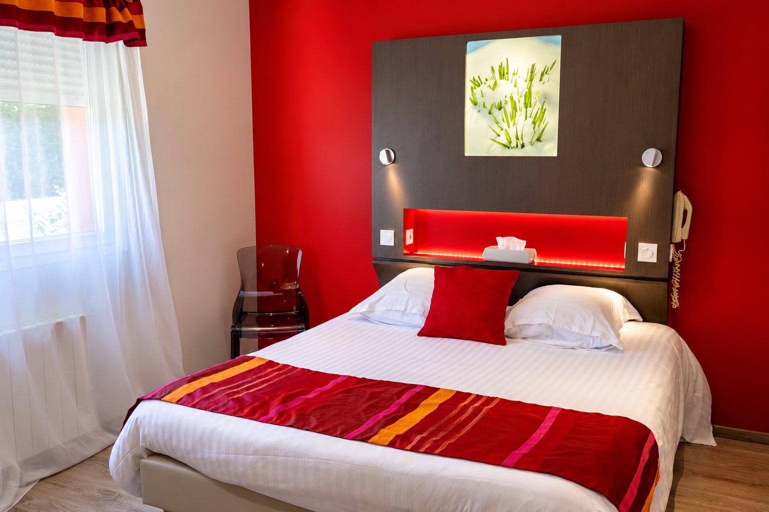 Chambre d’hôtel aux murs rouges avec un lit double habillé de blanc, un chemin de lit rayé rouge et orange, une tête de lit grise avec niche éclairée et un rideau voilage à la fenêtre.