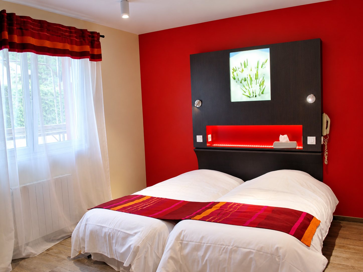 Chambre d’hôtel avec deux lits jumeaux aux draps blancs, un mur rouge, une tête de lit sombre avec niche lumineuse et un rideau blanc à la fenêtre.