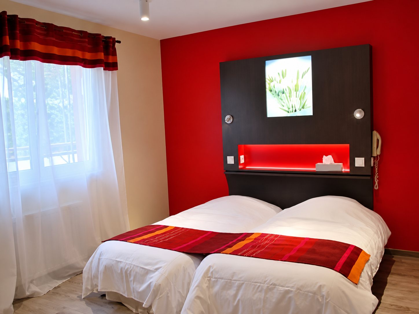 Une chambre d’hôtel lumineuse avec un mur rouge, une tête de lit sombre avec tableau et deux lits jumelés habillés de blanc et de chemins de lit rayés rouge et orange.