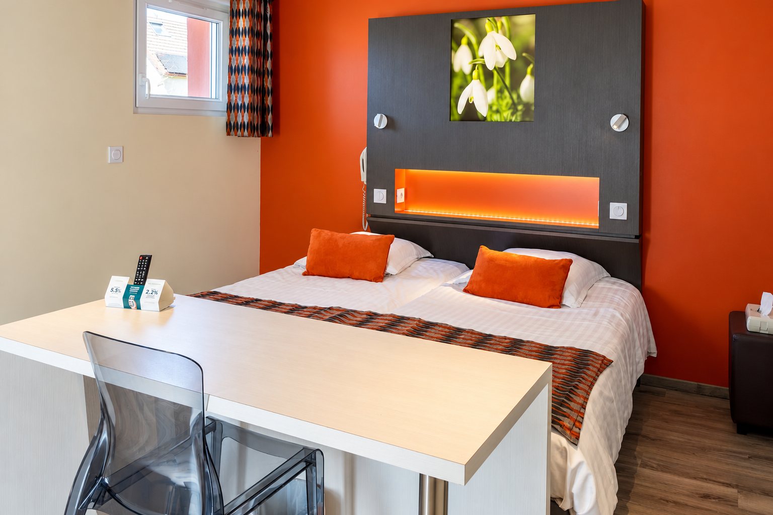 Chambre d’hôtel aux murs orange avec deux lits réunis, une tête de lit grise ornée d’une photo de perce-neige et un bureau blanc au premier plan.