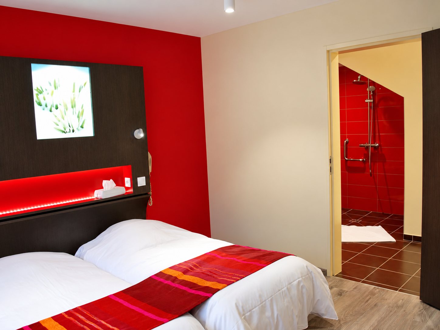 Chambre d’hôtel moderne aux murs rouges avec deux lits blancs ornés d’un chemin de lit rayé et une salle de douche carrelée de rouge en arrière-plan.