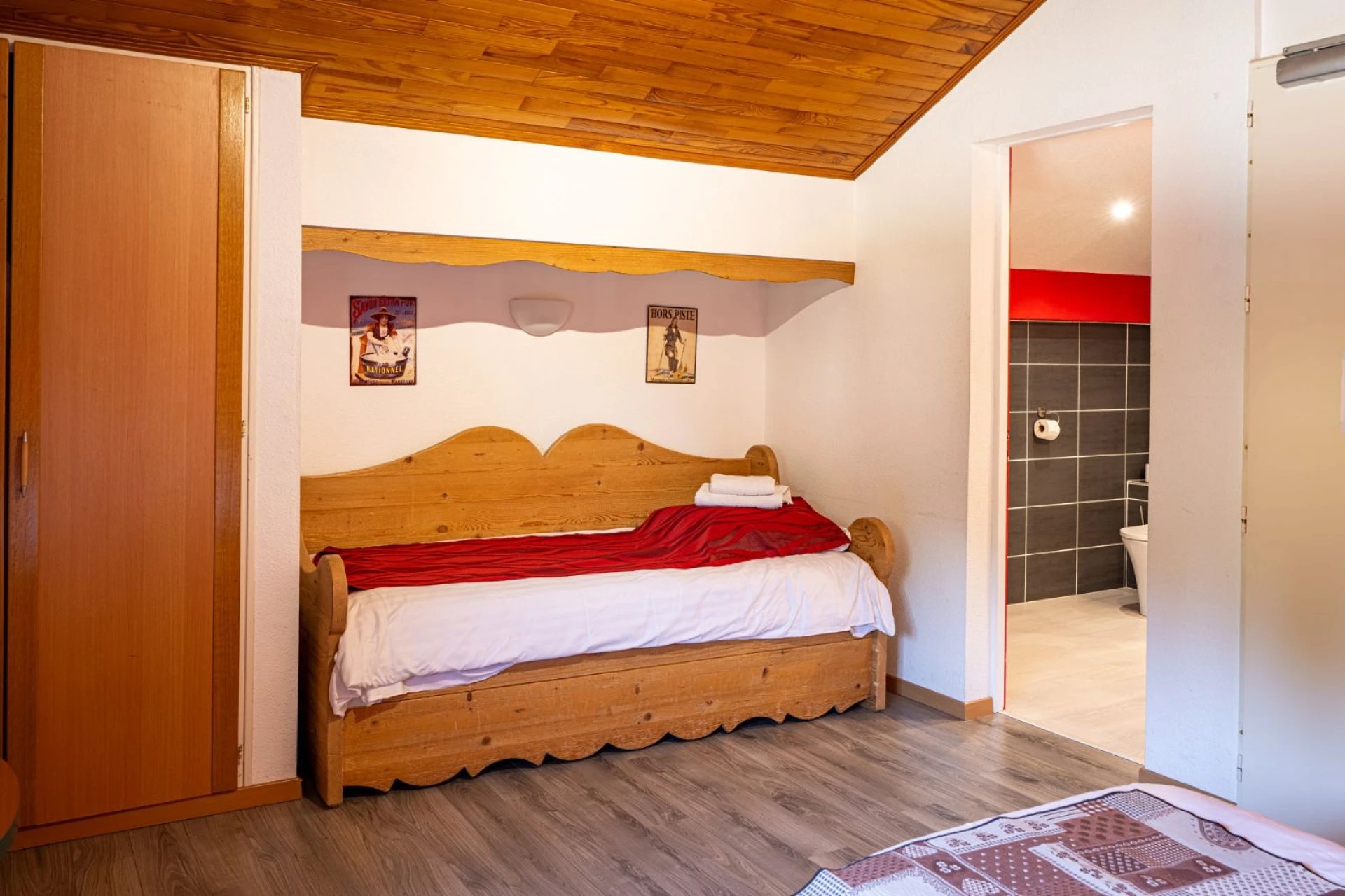 Chambre sous les combles aux finitions en bois avec un lit banquette sculpté, des affiches de ski au mur et une salle de bain carrelée en gris visible à droite.