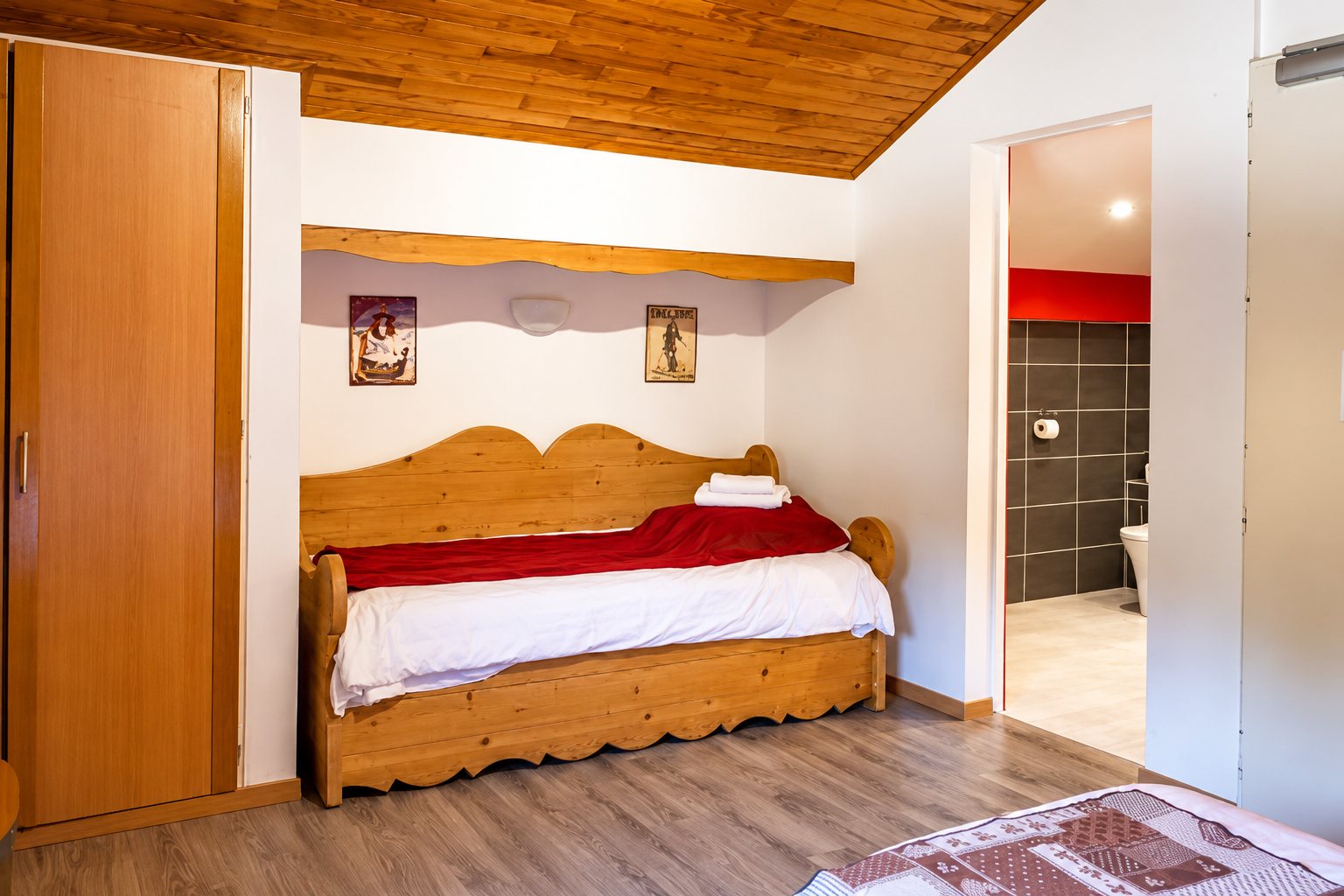 Chambre sous les combles avec un lit banquette en bois clair recouvert de rouge et de blanc, une armoire et une salle de bain carrelée en gris visible par la porte ouverte.