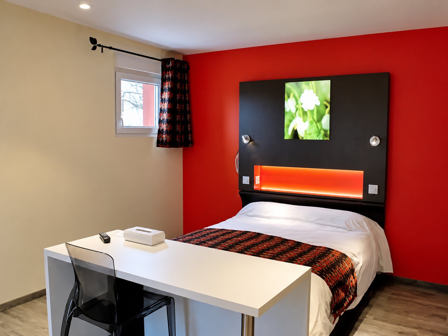 Chambre d’hôtel moderne avec un mur rouge, un lit double à tête de lit sombre éclairée en orange et un bureau blanc au premier plan.