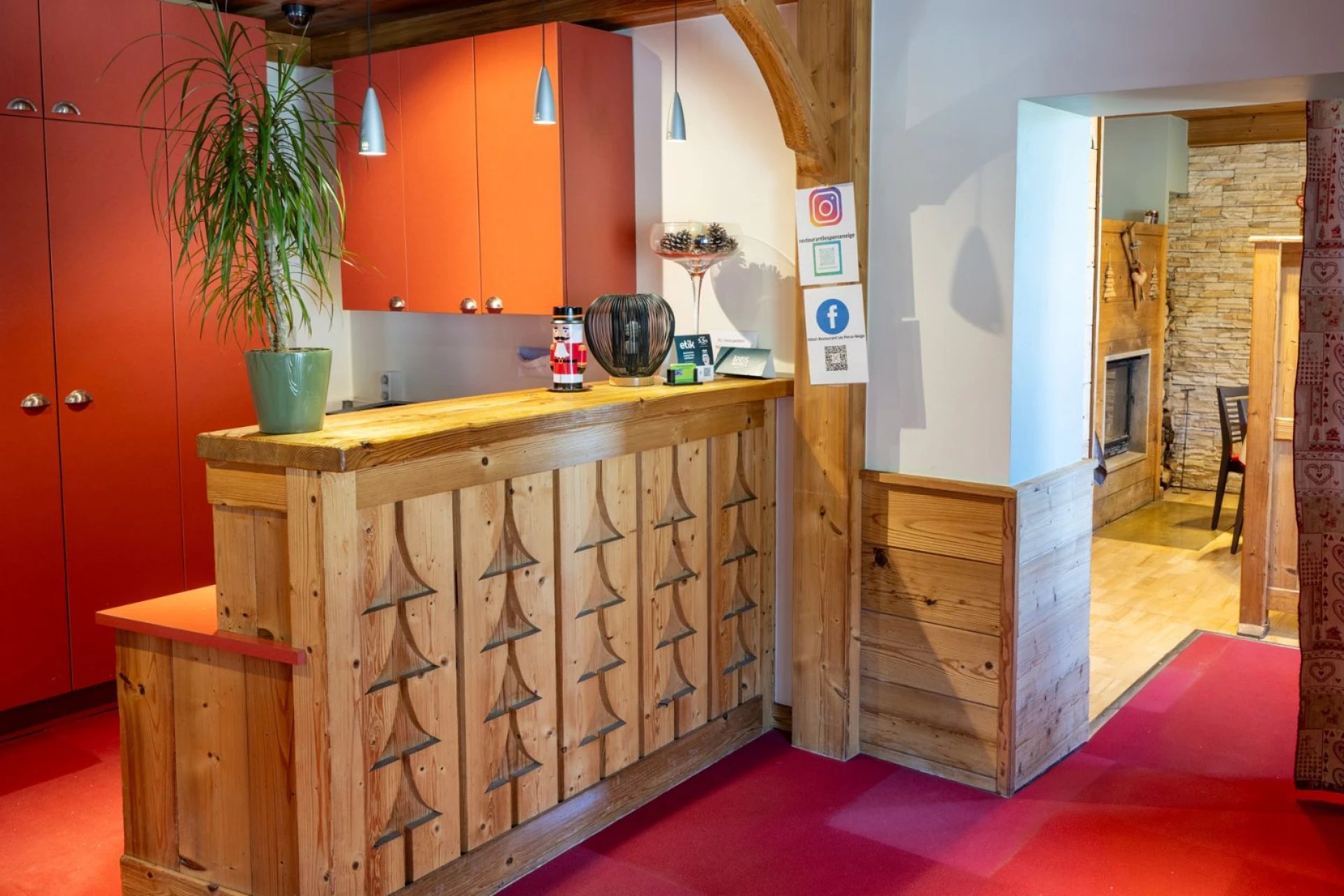 Un comptoir d’accueil en bois clair orné de sapins sculptés, devant des placards orange et un salon avec cheminée en arrière-plan.