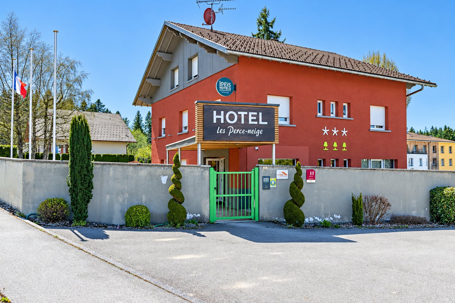 Façade d’un hôtel au crépi rouge portant l’enseigne Hotel les Perce-neige, avec un portail vert et des buis taillés en spirale devant le bâtiment.