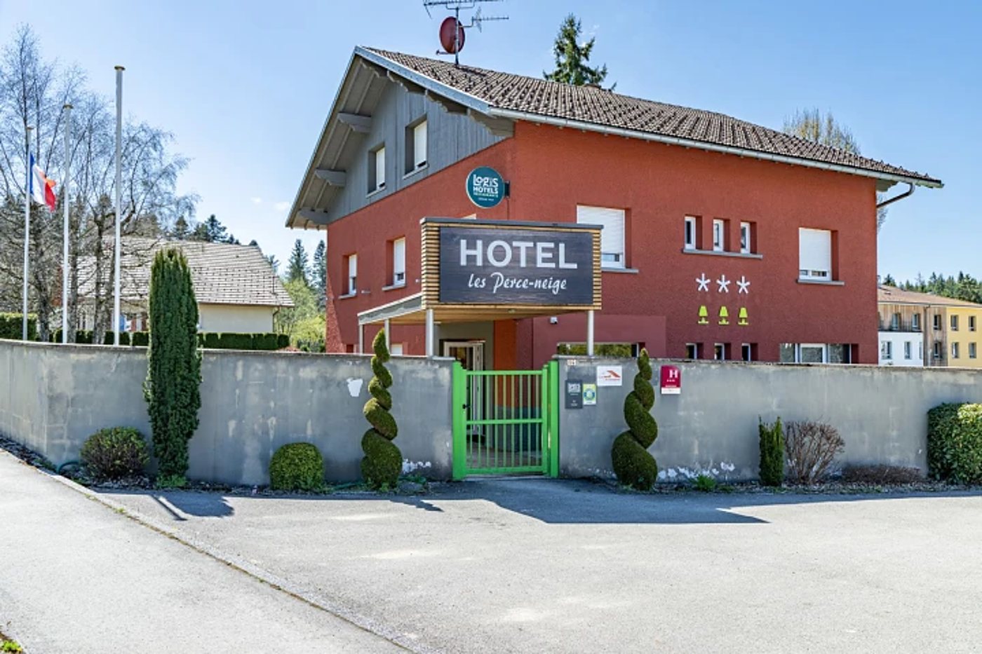 Façade rouge d’un hôtel trois étoiles nommé Les Perce-neige, avec une enseigne en bois, un portail vert et des buis taillés en spirale devant un mur gris.