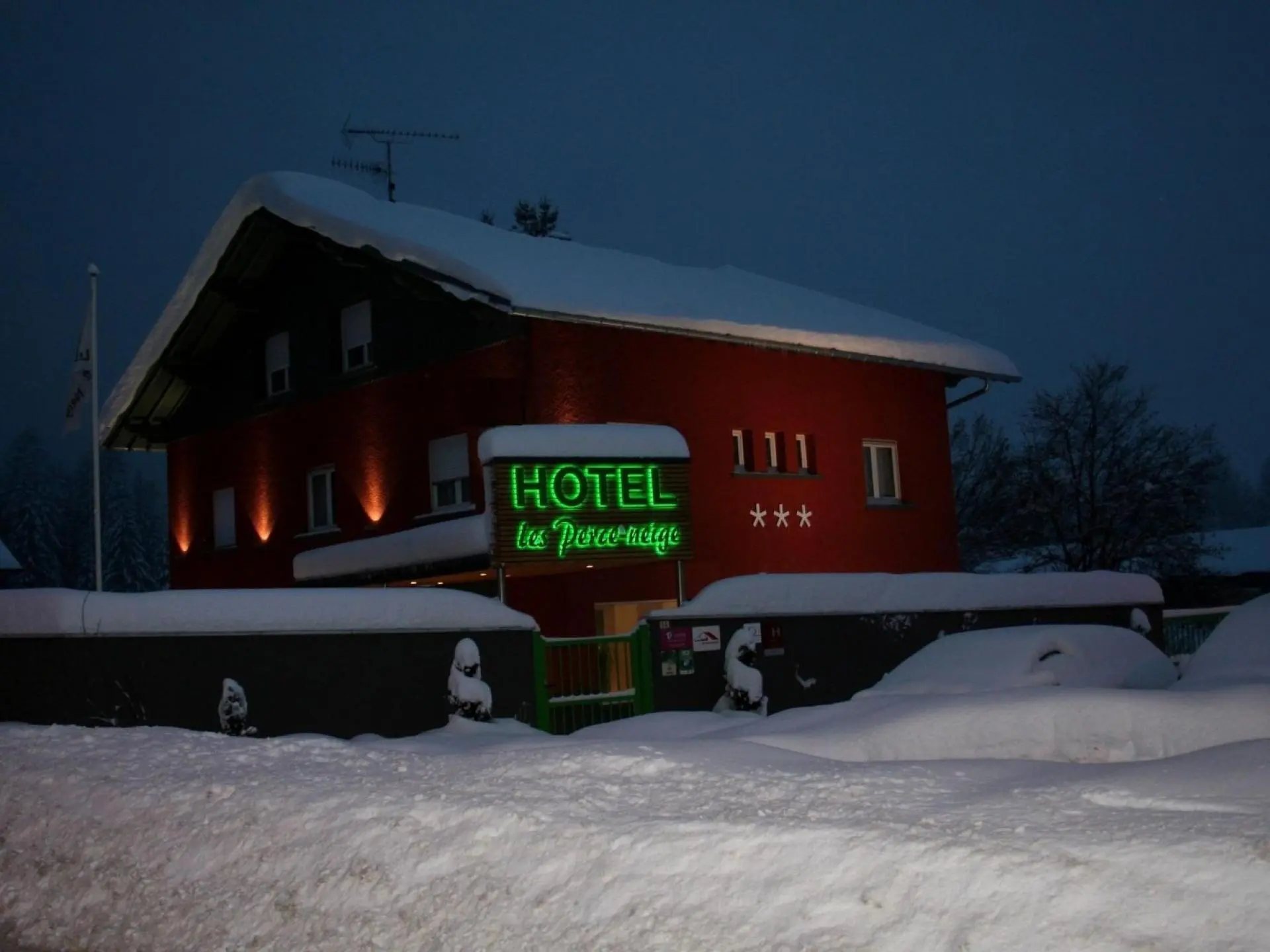 Façade d’un hôtel rouge trois étoiles sous la neige, de nuit, avec une enseigne lumineuse verte au néon.