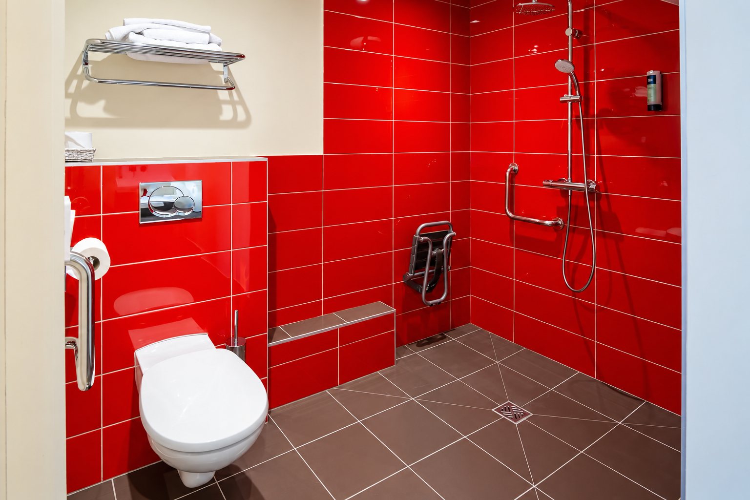 Salle de bain adaptée aux personnes à mobilité réduite avec murs en carrelage rouge, barres d’appui, siège de douche rabattable et WC suspendu.