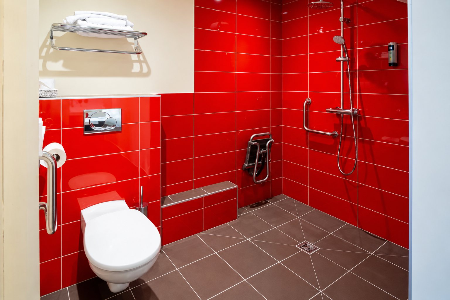 Salle de bain adaptée aux personnes à mobilité réduite, avec murs en carrelage rouge, WC suspendu, barres d’appui et douche avec siège rabattable.