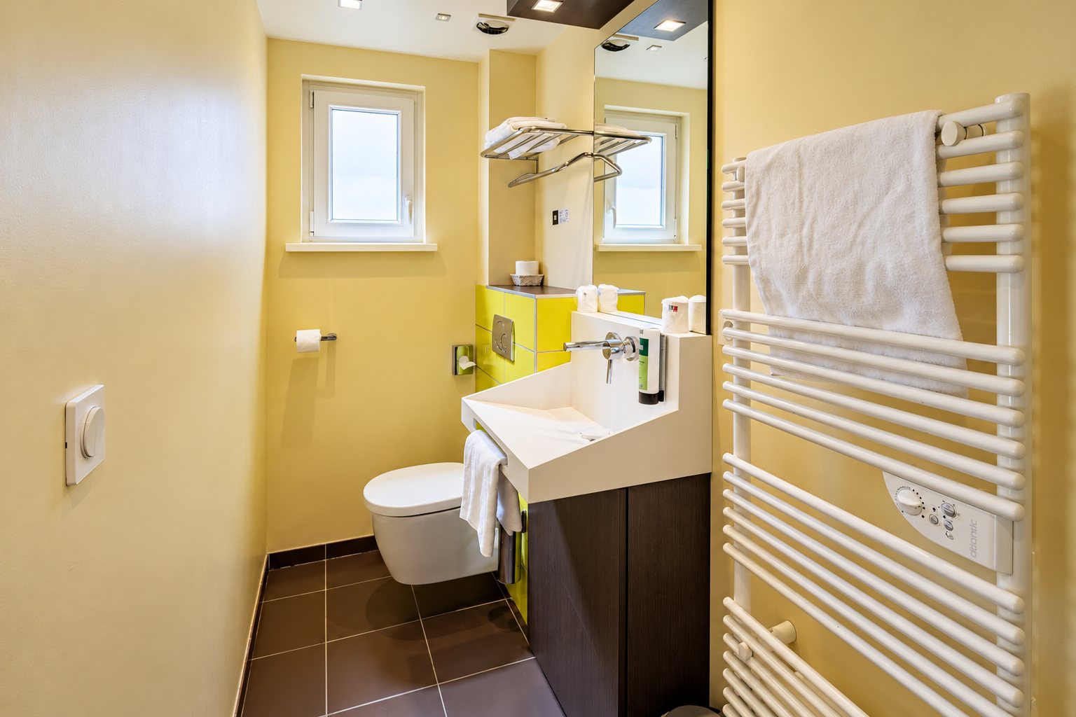 Salle de bain aux murs jaunes avec WC suspendu, lavabo blanc sur meuble brun, miroir et radiateur sèche-serviettes avec une serviette blanche.