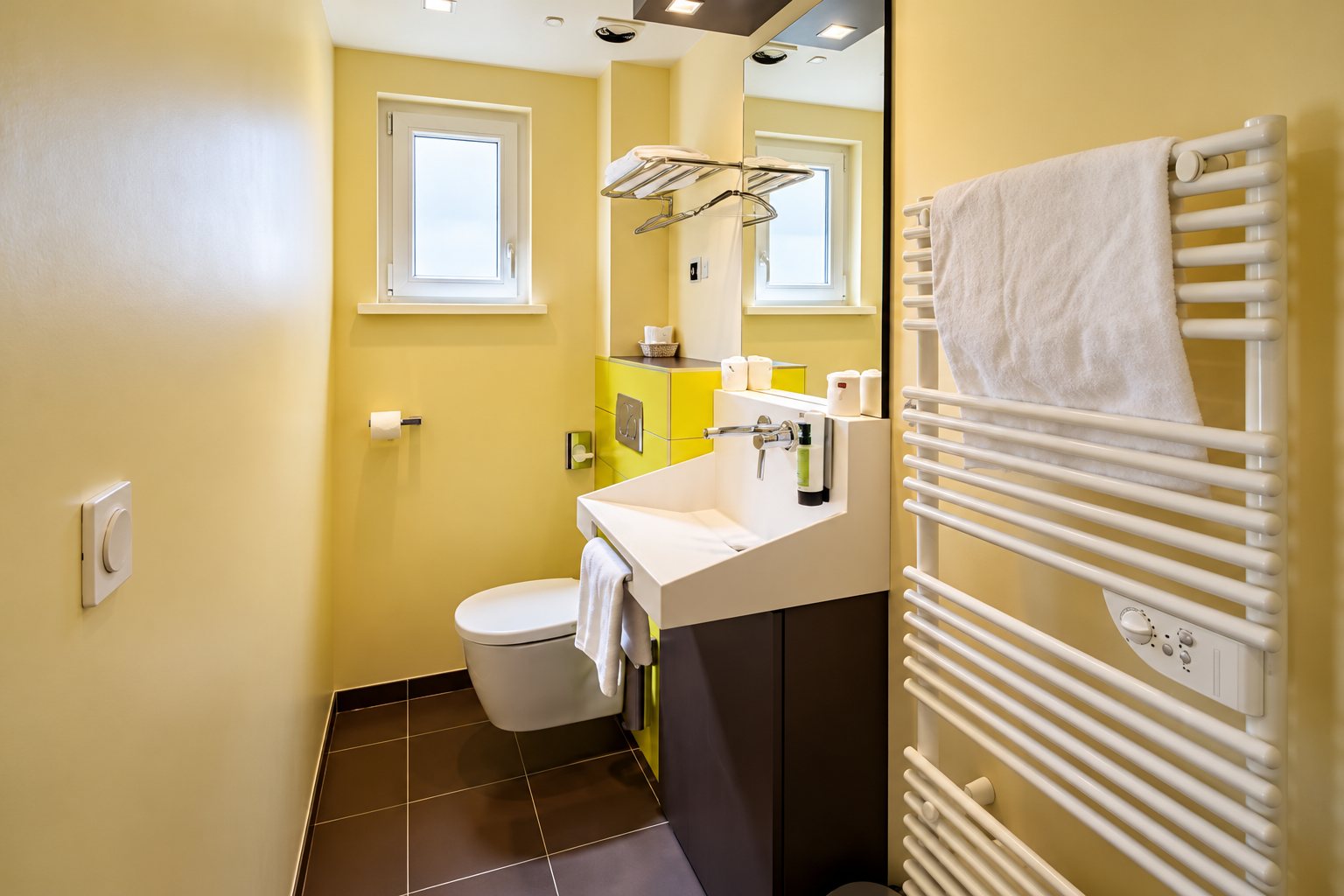 Petite salle de bain aux murs jaune clair avec lavabo suspendu, WC suspendu, miroir et radiateur sèche-serviettes blanc.