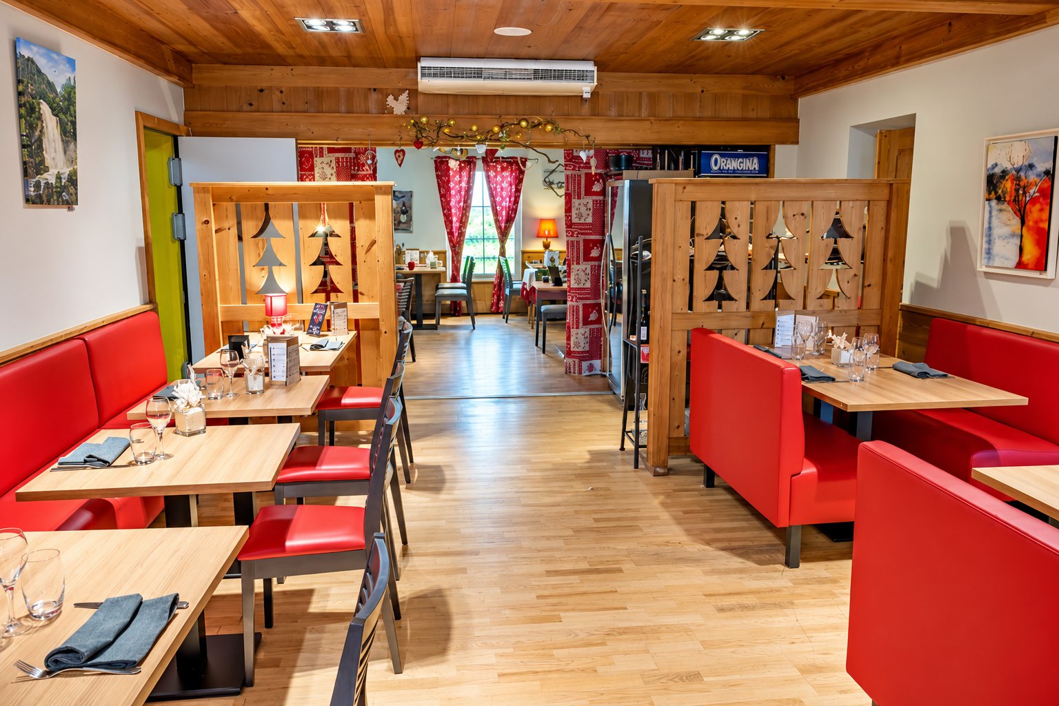 Intérieur d’un restaurant de style chalet avec banquettes rouges, tables en bois clair, cloisons ajourées à motifs de sapins et plafond en bois.