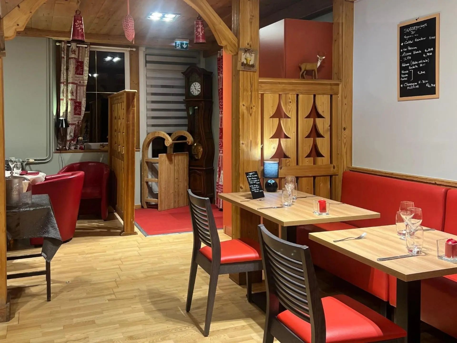 Intérieur d’un restaurant de style montagnard avec boiseries, banquettes et chaises rouges, tables dressées, horloge comtoise et ardoise de suggestions au mur.