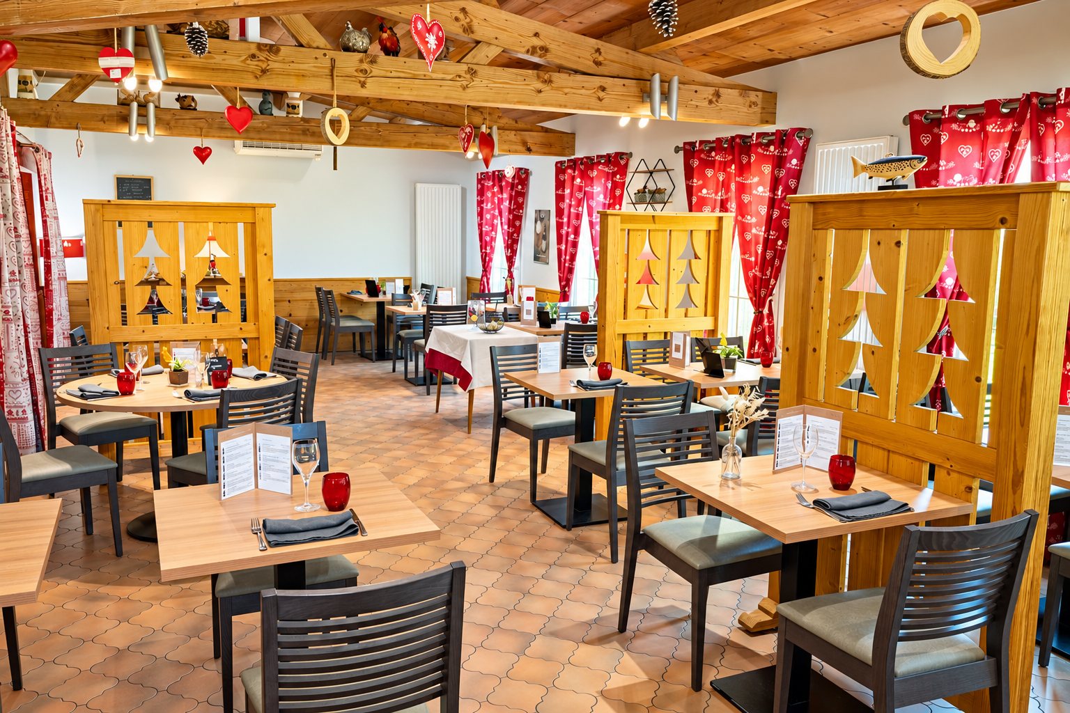 Salle de restaurant au décor montagnard avec tables en bois, chaises grises, cloisons en bois découpées en forme de sapins et rideaux rouges à motifs de cœurs.