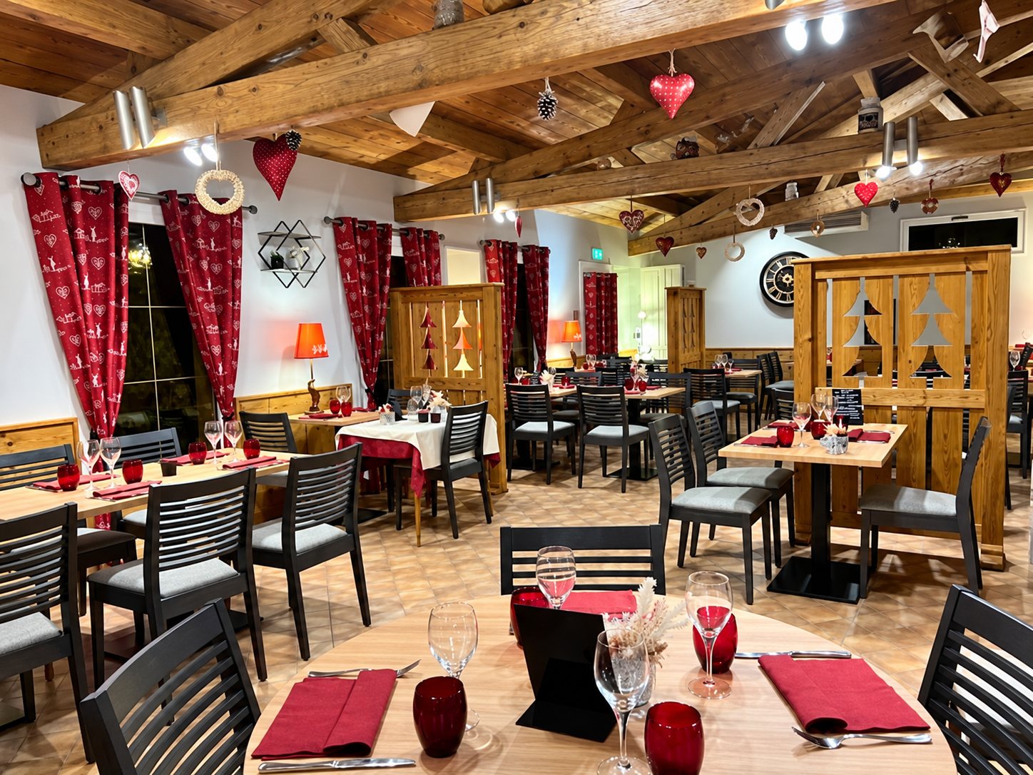 Salle de restaurant de style chalet avec poutres en bois, rideaux rouges, tables dressées avec serviettes et verres rouges.