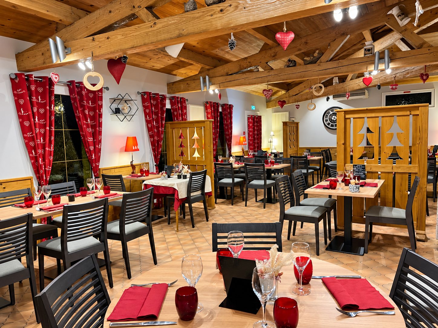 Salle de restaurant de style chalet avec poutres en bois, rideaux rouges à motifs de cœurs et tables dressées avec serviettes et verres rouges.