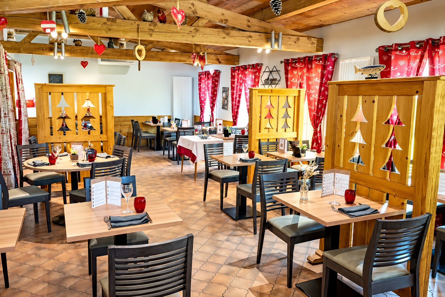 Salle de restaurant rustique aux tables en bois dressées, séparées par des cloisons en bois décorées de sapins et ornées de rideaux rouges.
