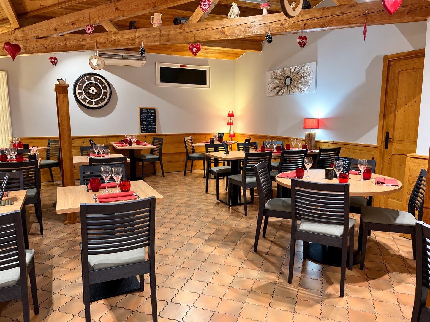 Salle de restaurant au décor montagnard avec poutres en bois, cœurs rouges suspendus, tables rondes et carrées dressées avec verres et serviettes rouges.