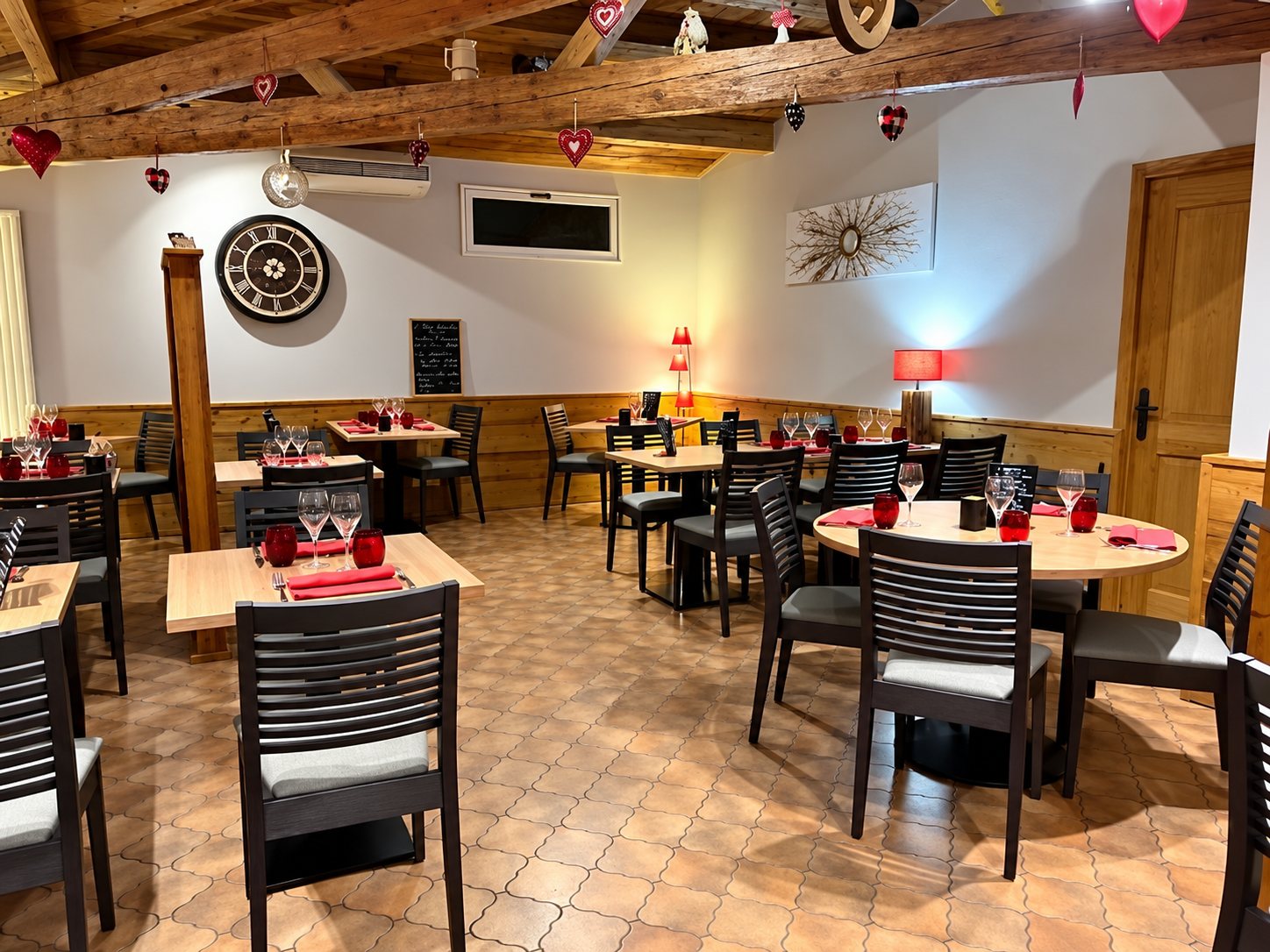 Salle de restaurant aux poutres apparentes, avec tables en bois dressées, chaises sombres, grande horloge murale et petits cœurs rouges suspendus au plafond.