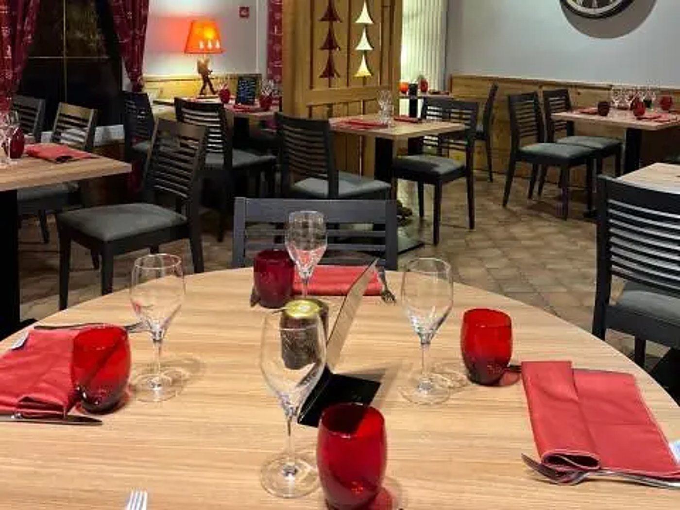 Salle de restaurant au décor montagnard avec tables en bois dressées de verres, de gobelets et de serviettes rouges.
