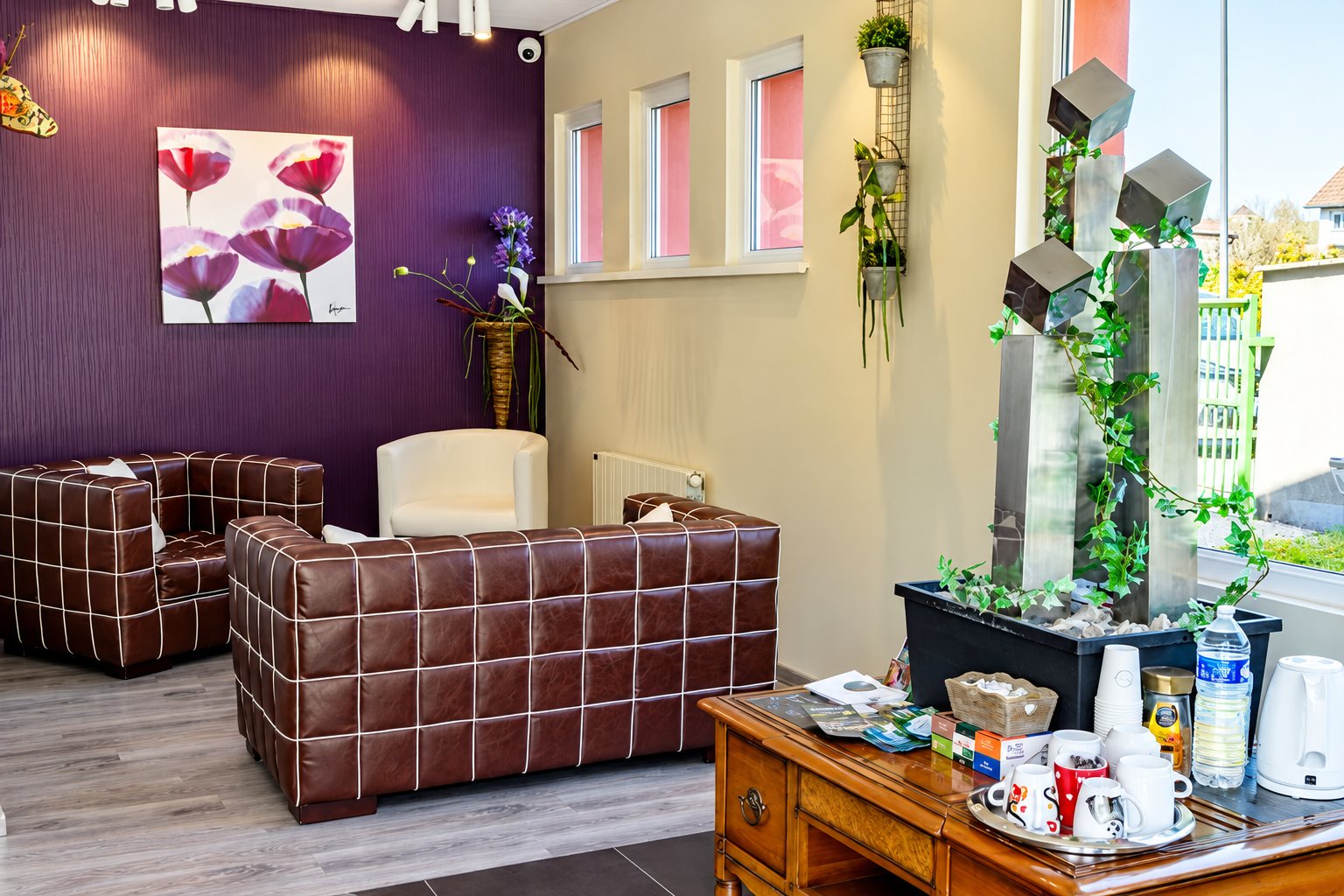 Salon d’accueil lumineux avec canapés en cuir brun, mur violet orné de coquelicots et table proposant tasses, bouilloire et bouteille d’eau près d’une fontaine décorative.