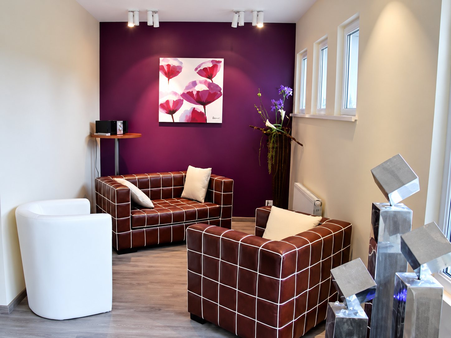 Un espace d’exposition lumineux avec deux canapés en cuir bordeaux à carreaux, un fauteuil blanc, un mur violet orné d’un tableau de fleurs et des luminaires design en métal au premier plan.