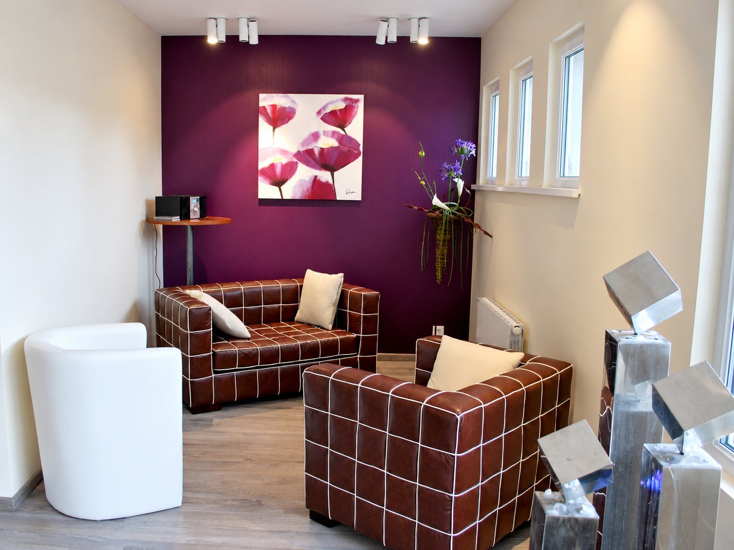 Un salon lumineux aux canapés en cuir brun quadrillé de blanc, avec un mur violet orné d’un tableau de coquelicots et des sculptures métalliques au premier plan.